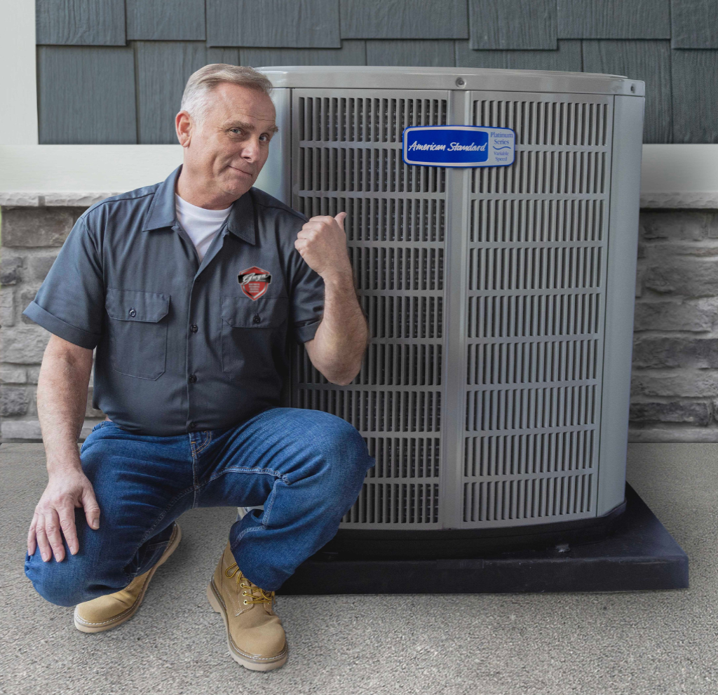 Man kneeling beside an outdoor air conditioner unit, smiling and pointing at it