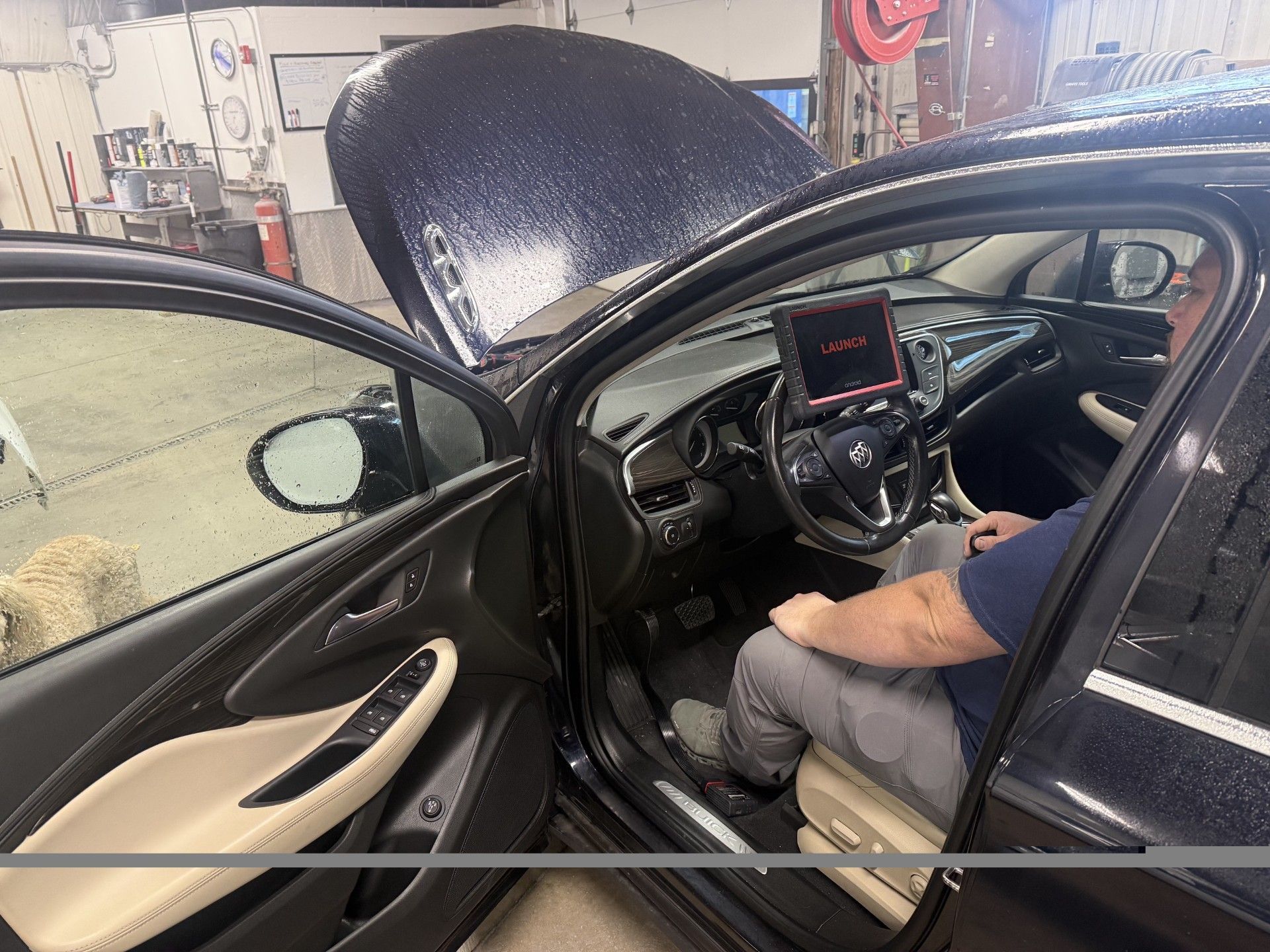 Car interior with hood open; person seated in driver’s seat using a laptop diagnostic tool.