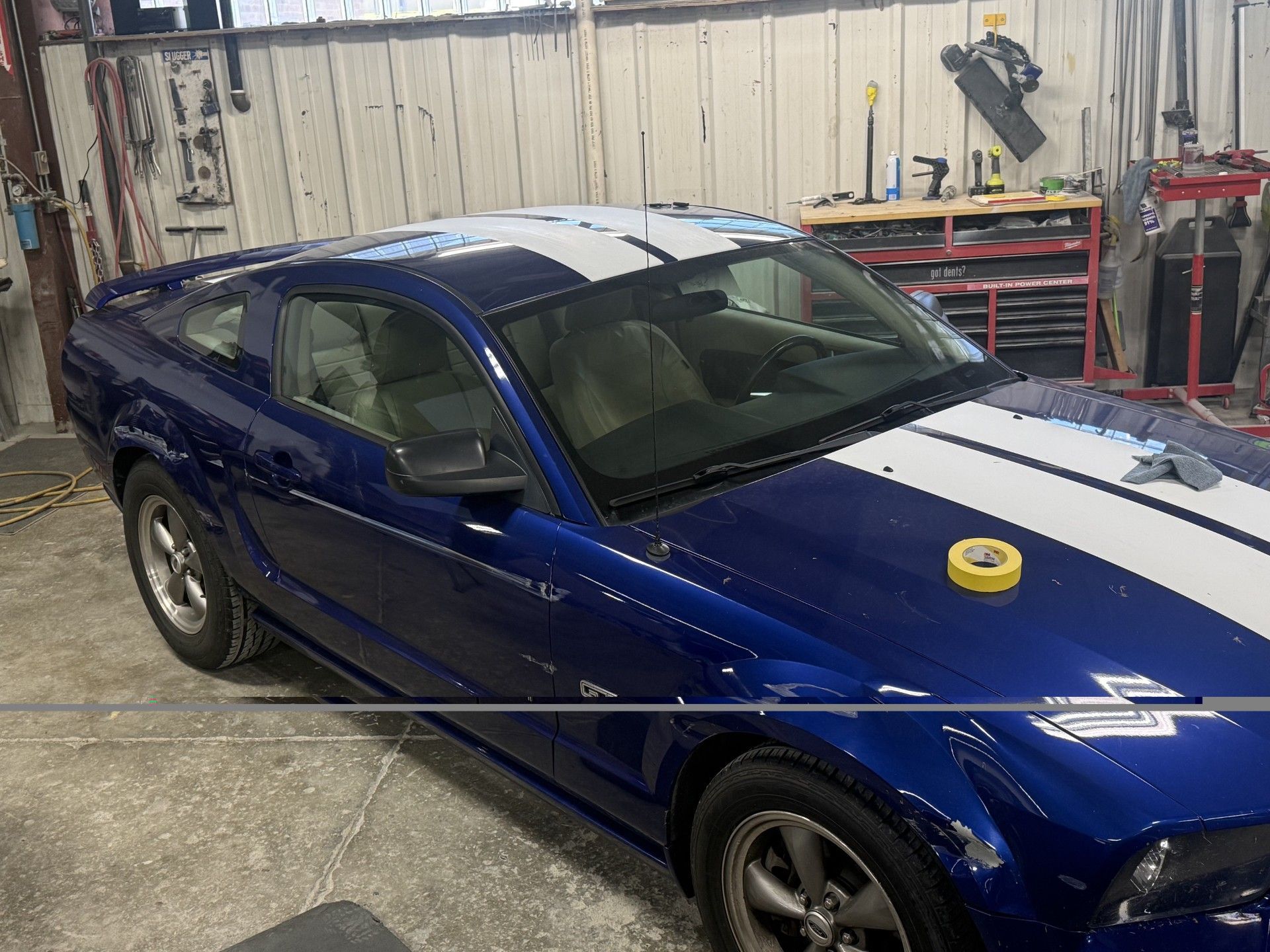 Blue sports coupe in an auto shop, parked indoors with a yellow tape roll on the hood.