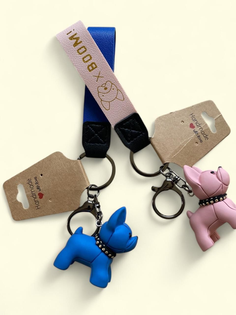 Dos llaveros con forma de perro, con correas a rayas y etiquetas colgantes, uno azul y otro rosa, sobre un fondo claro.