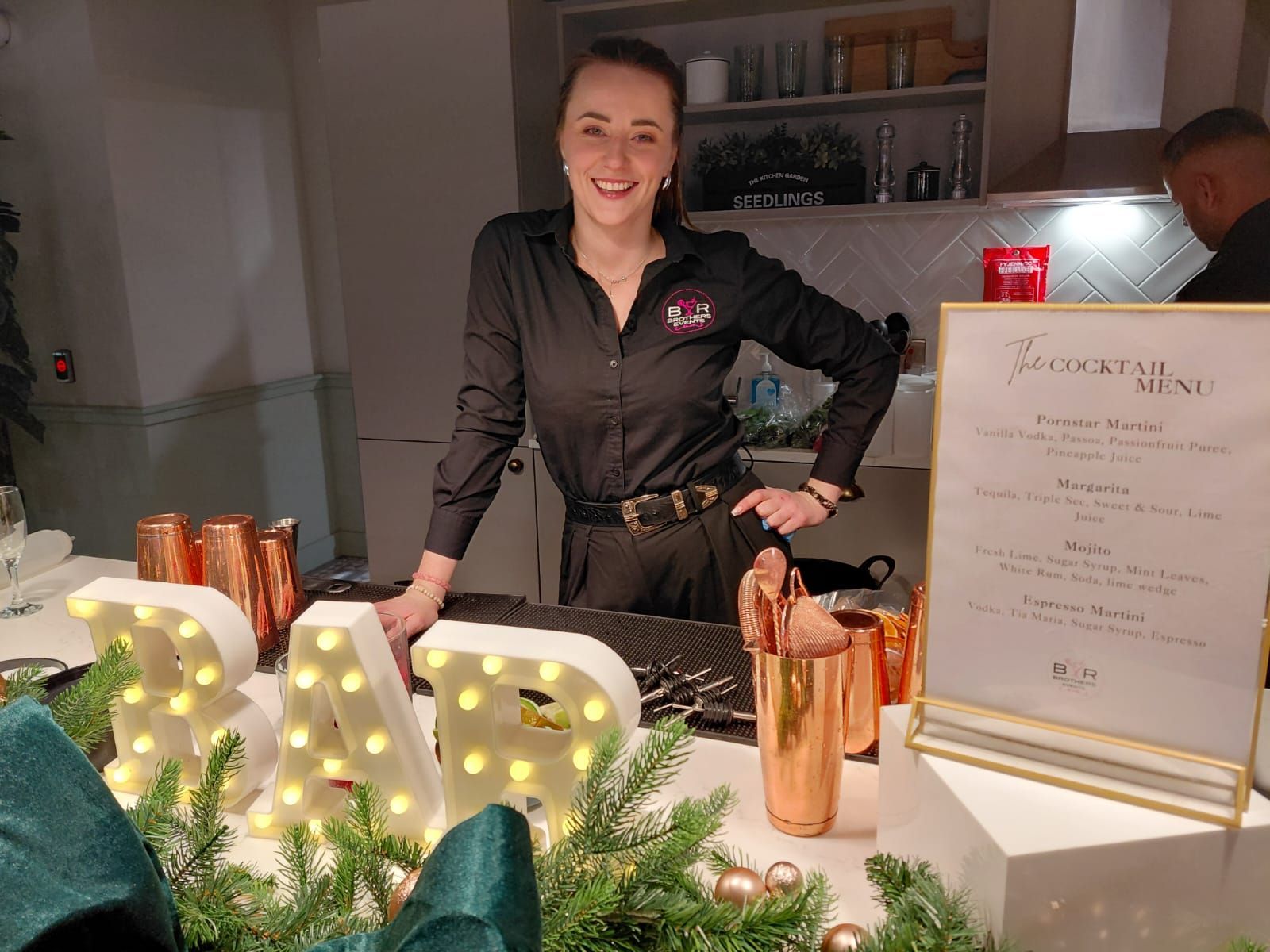 Woman behind a decorated bar with lights, candles, and a menu sign at an indoor event