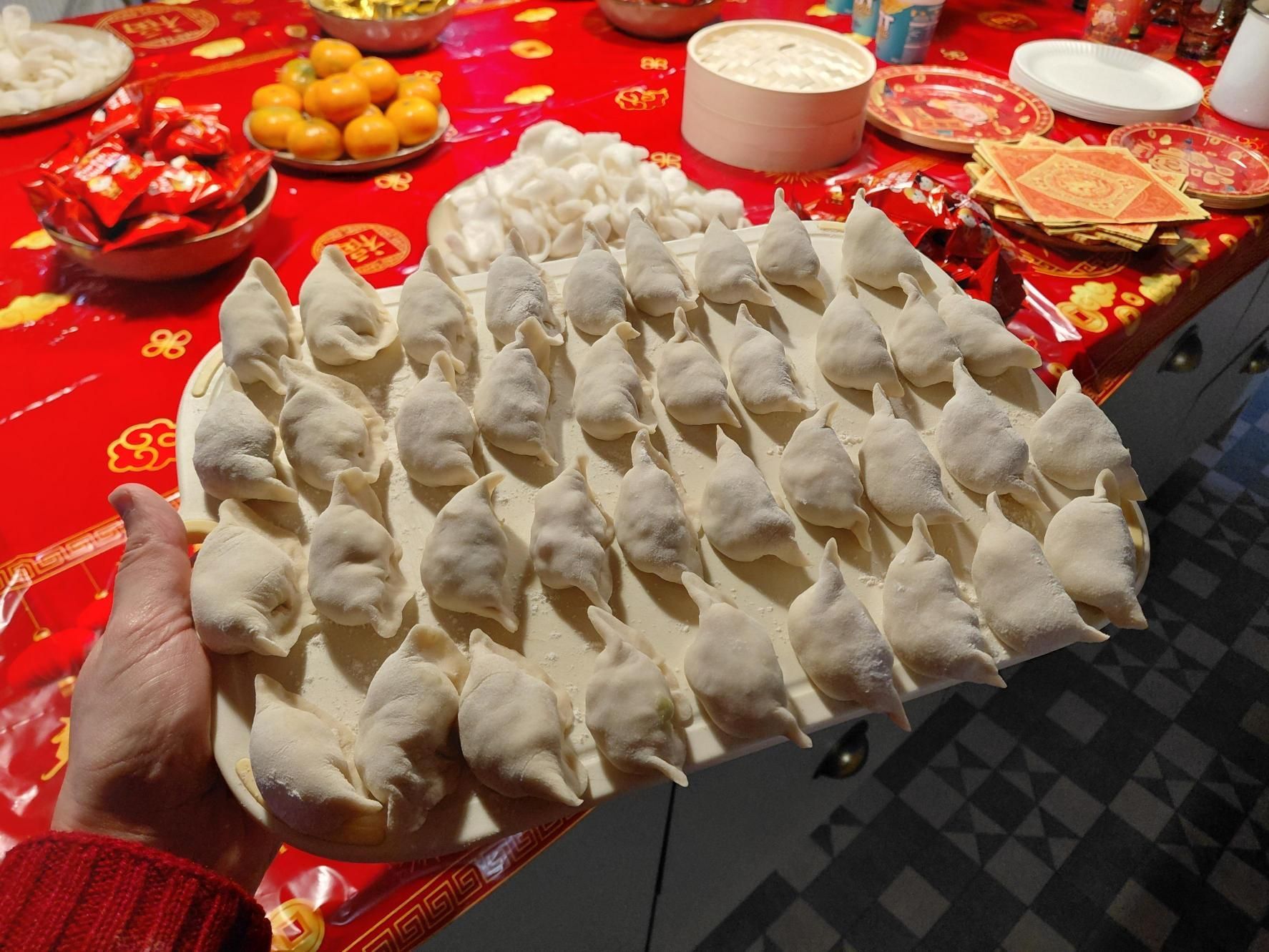 Hand holding a tray of white dumplings over a red tablecloth with fruit and dishes.