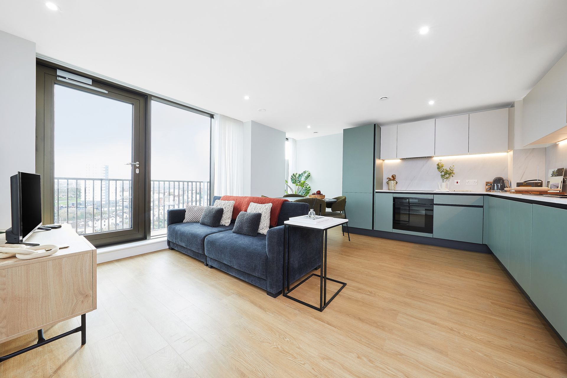 Bright open-plan living room with blue sofa, wooden floor, and teal kitchen cabinetry