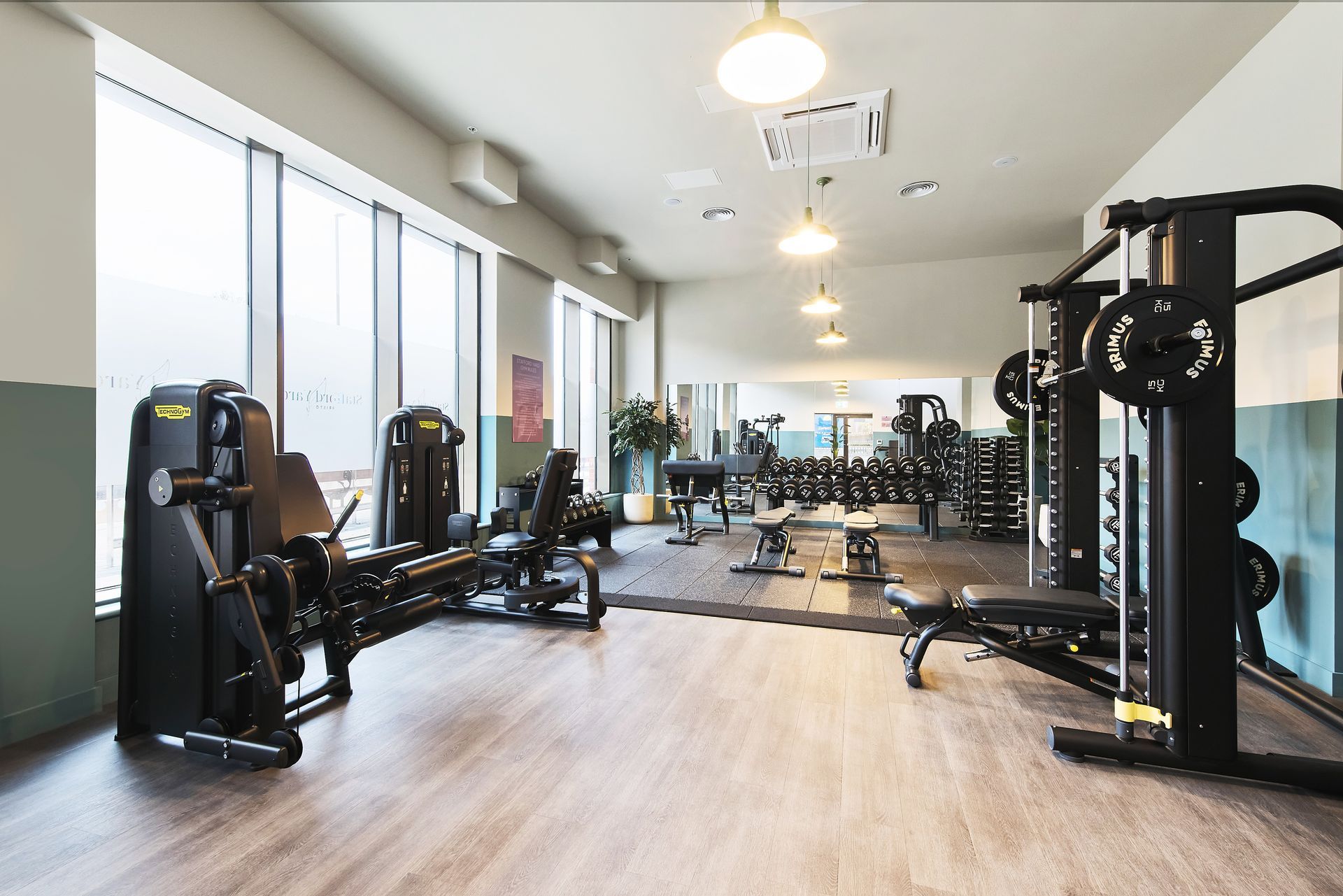 Gym with exercise machines, mirrors, and large windows letting in daylight.