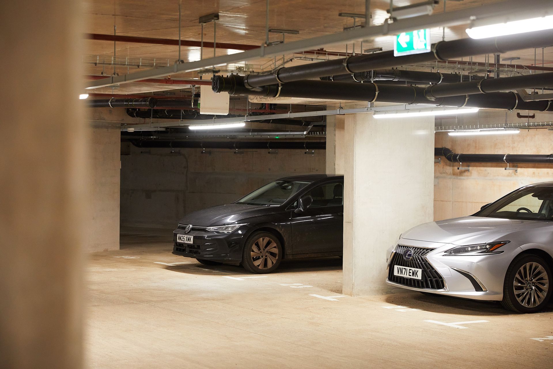 Underground parking garage with two parked cars, concrete pillars, and fluorescent lighting.
