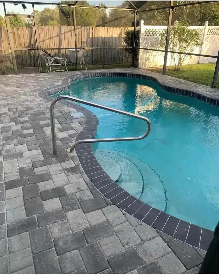 Backyard swimming pool with curved silver handrail and gray paver deck, bordered by a wooden fence.