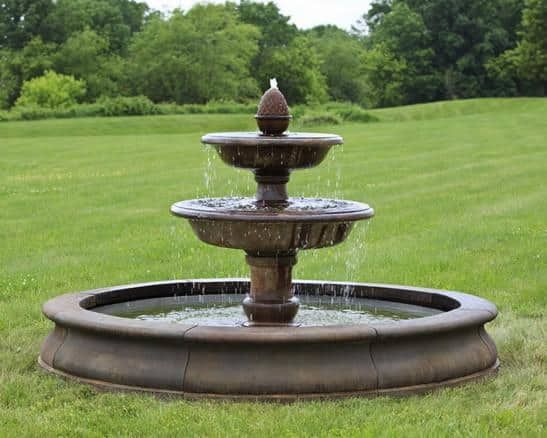 Three-tier stone fountain spraying water on a grassy lawn with trees in the background