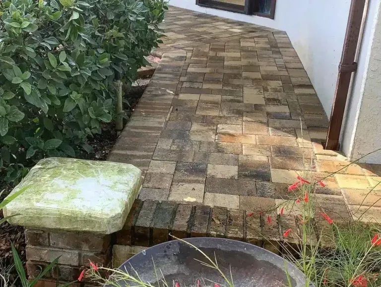 Brick patio walkway beside a white house, bordered by green shrubs and a small stone bench.