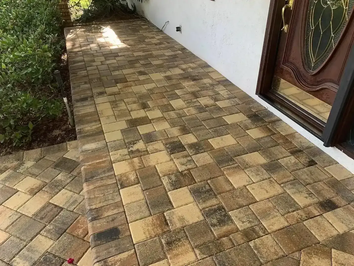Tiled outdoor walkway beside a white wall and wooden door, with shrubbery along the left side.