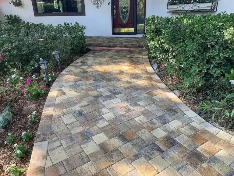 Brick walkway leading to a house entrance, bordered by green shrubs and flower beds.