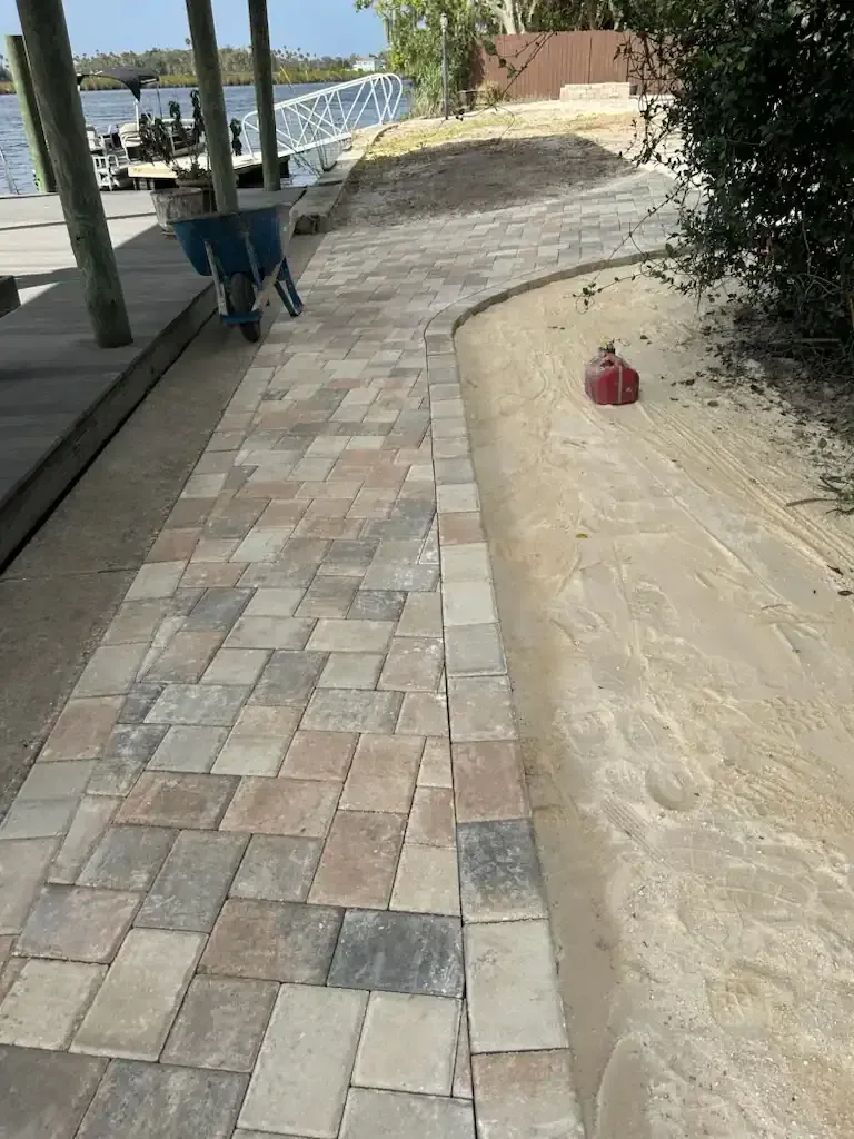 Paved walkway beside water, with a blue wheelbarrow and a small red object on the sand.