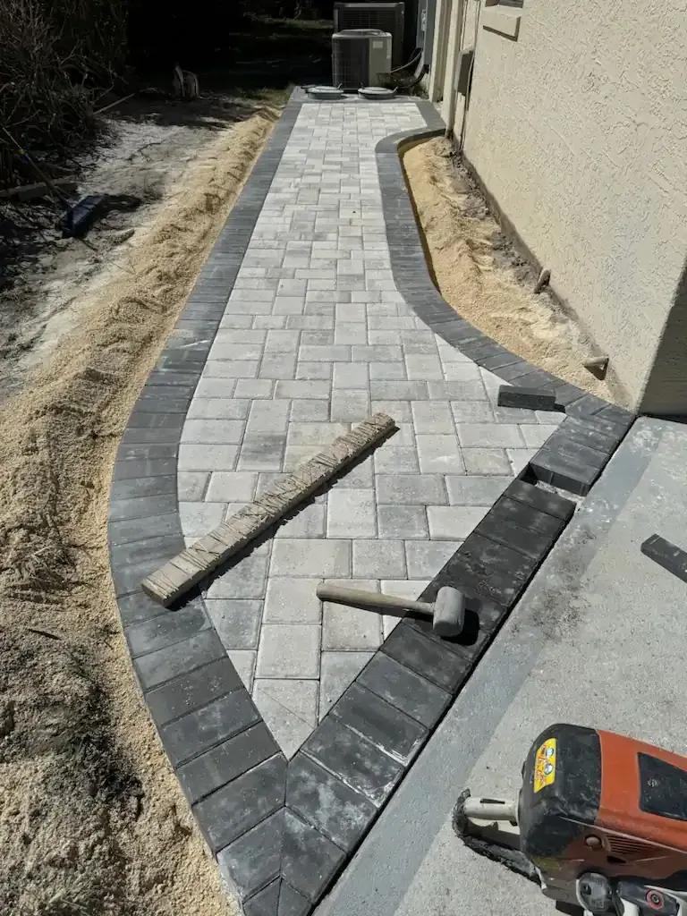 New paver walkway under construction beside a beige wall, with tools and edging stones visible.