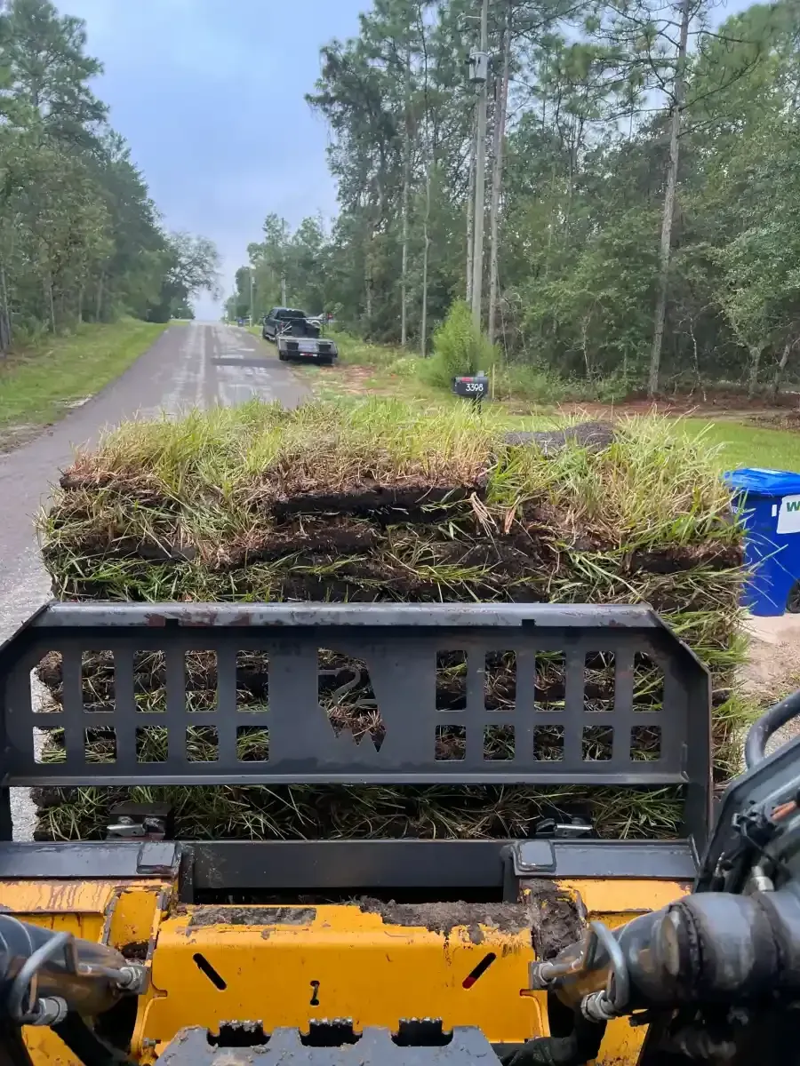 View from a yellow off-road vehicle carrying brush on a rural gravel road with trees and a parked car ahead