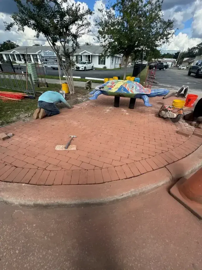 Workers repairing a brick plaza under trees, with tools, materials, and houses in the background