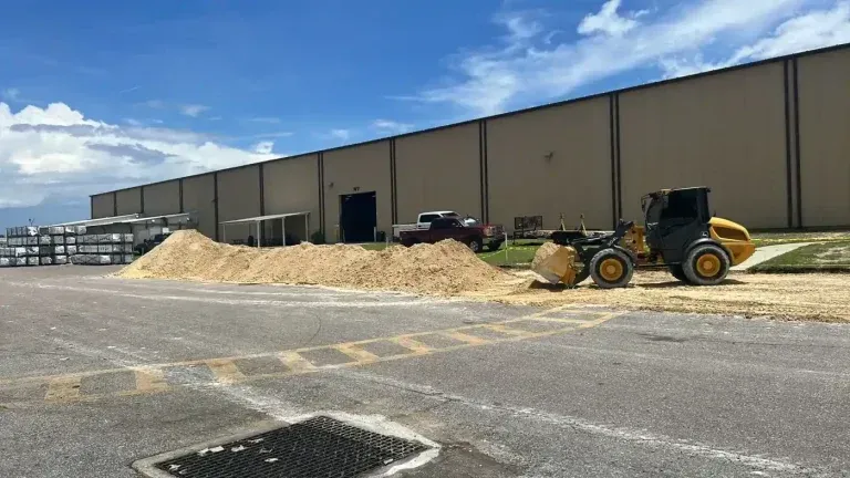 Construction site with tan warehouse, pile of gravel, and yellow loader under a partly cloudy sky