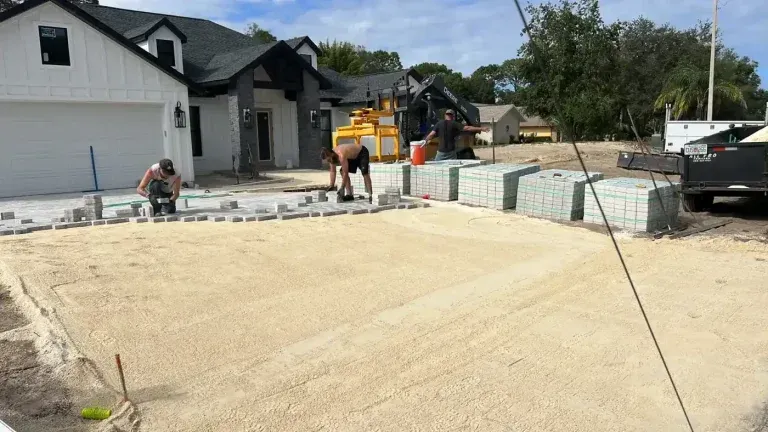 Workers paving a new concrete driveway beside a suburban house, with a truck and materials nearby.