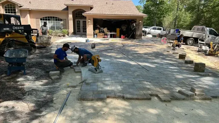 Workers laying stone pavers in a driveway beside a house, with construction equipment and vehicles nearby