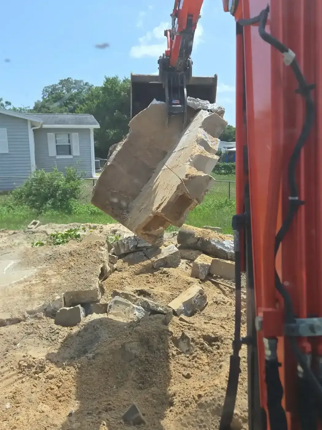 Excavator lifting broken concrete slabs at a demolition site beside a house.