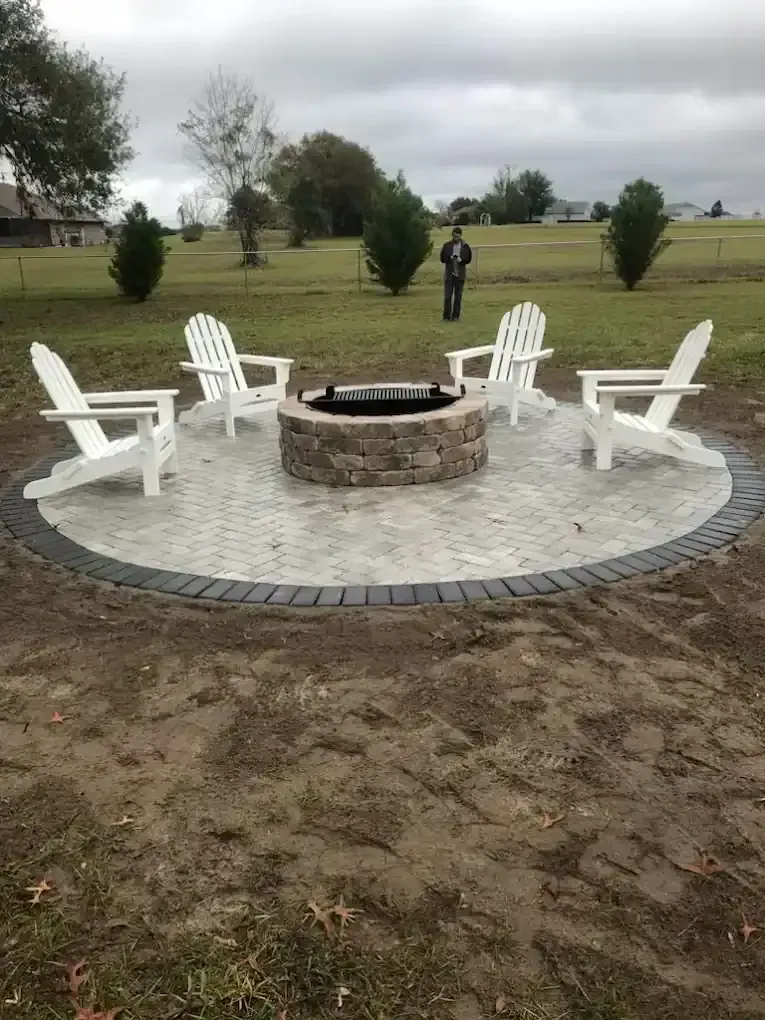 Stone fire pit with four white Adirondack chairs on a circular patio in a grassy park.