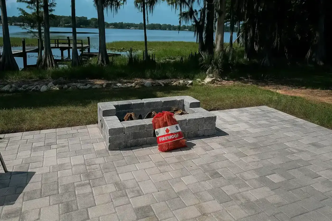 Stone fire pit on a paved patio with a red bag, overlooking a lake and trees.