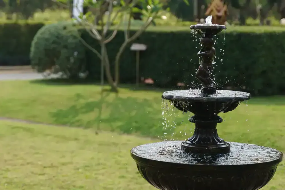 Black tiered garden fountain spraying water on a green lawn with hedges in the background
