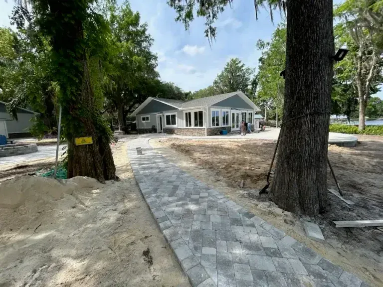 Stone walkway leading to a gray house under construction, framed by large trees and sandy ground