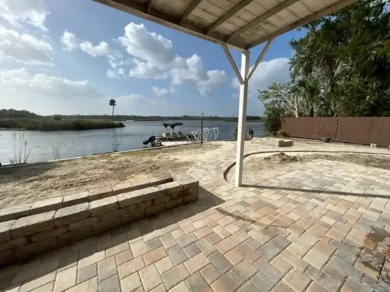 Covered riverside patio with brick pavers, steps, and a dock on calm water under a blue sky