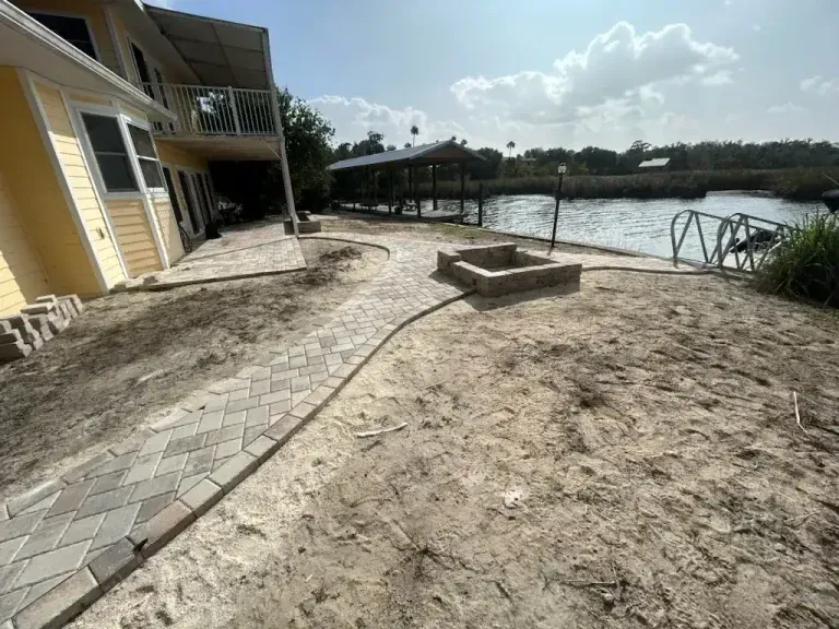 Sandy waterfront patio beside a yellow house, with a paved walkway and dock over the water.