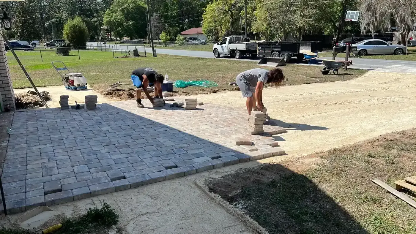 Workers laying a light-colored paver patio beside an existing dark stone walkway in a grassy yard.
