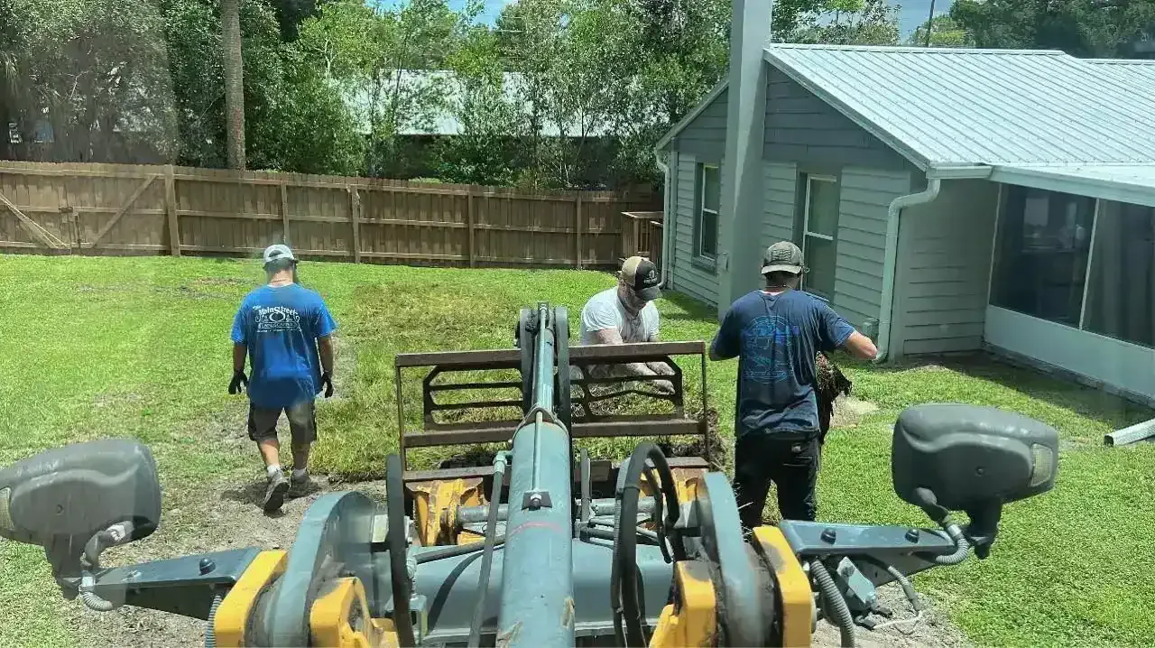 Two workers with a small excavator clearing gravel in a backyard beside a gray shed.