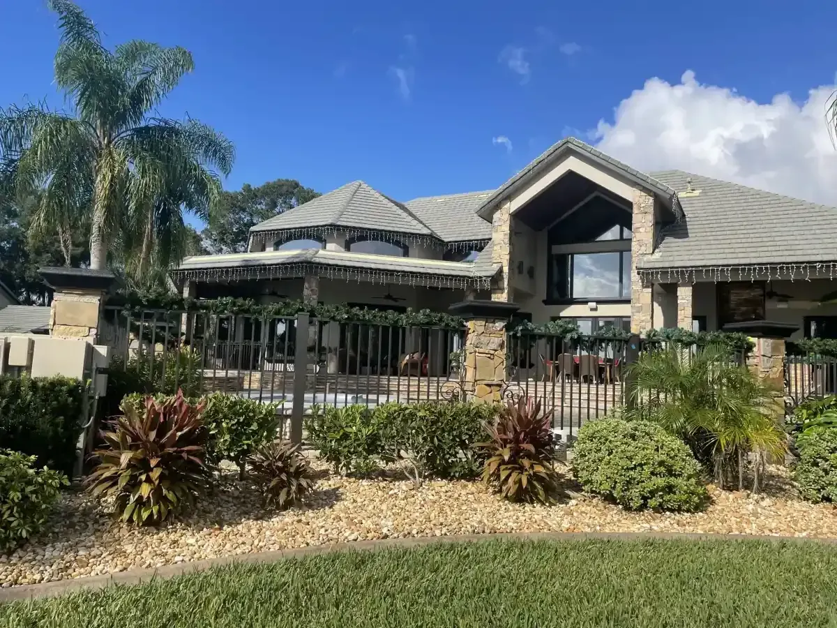Modern house with a gray roof, tropical landscaping, and a fenced yard under a blue sky.