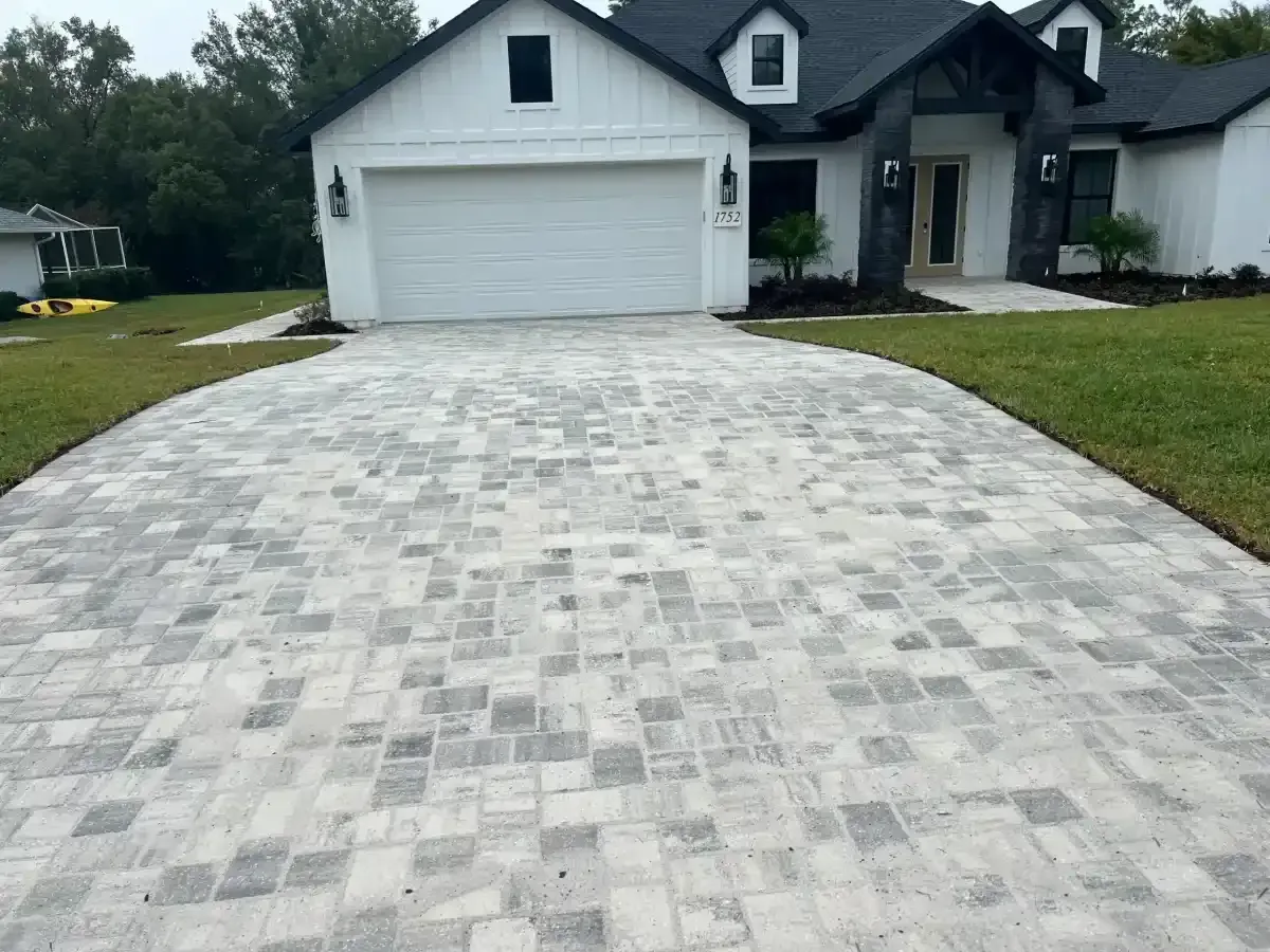 White house with a large light-gray paver driveway and attached garage, viewed from the front lawn.