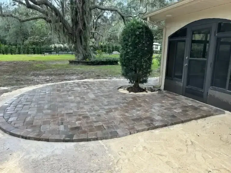 Brick patio beside screened porch and trimmed shrub in a grassy yard
