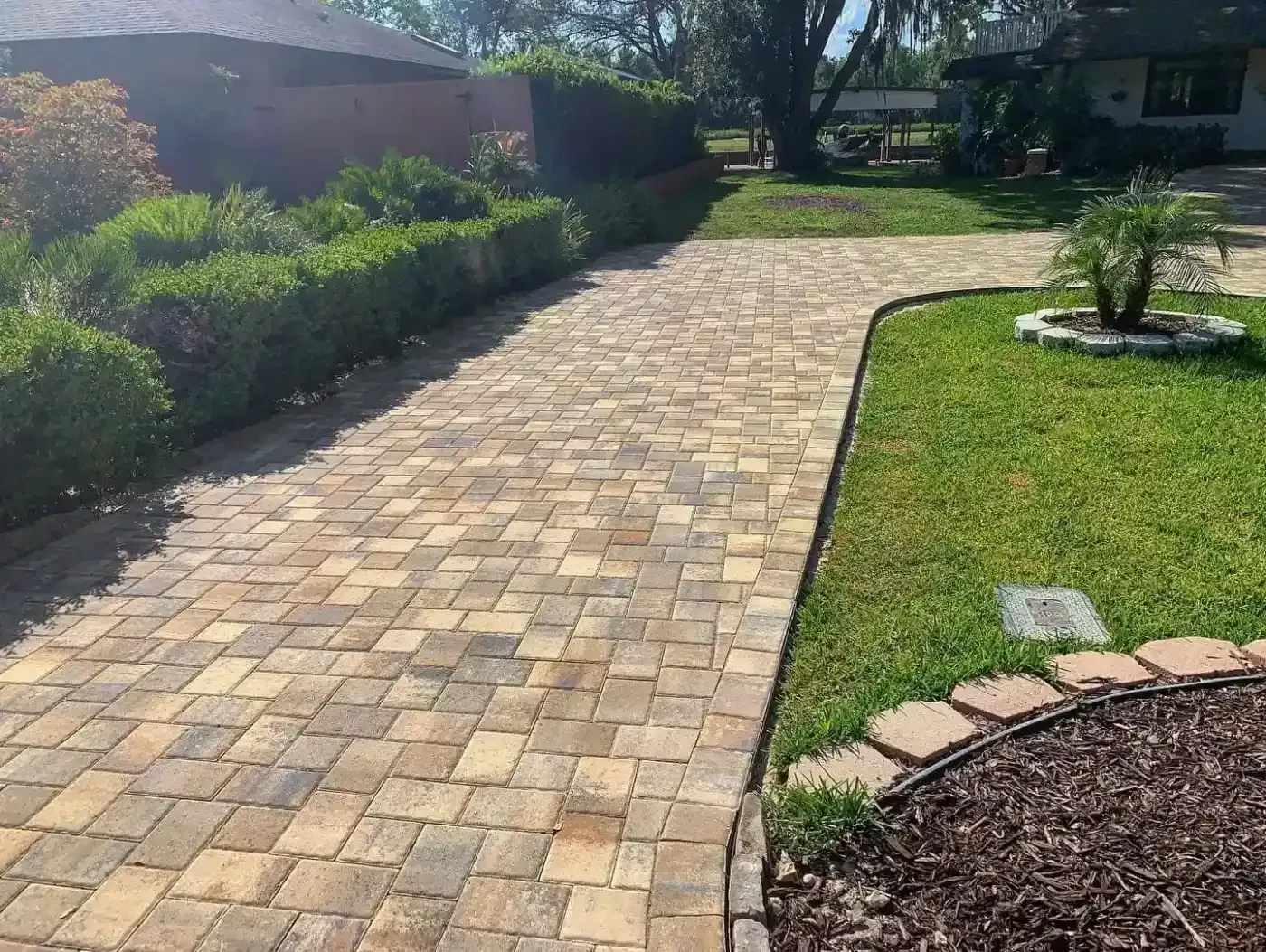 Brick-paved driveway curving past a lawn and garden beds beside a house in bright sunlight