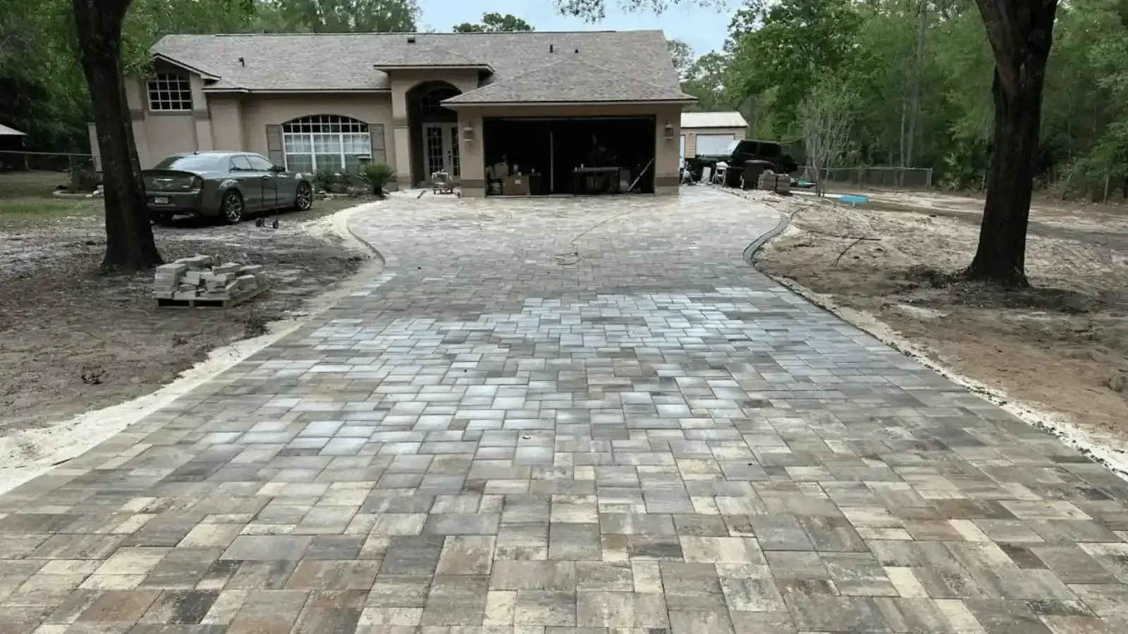 Wide stone driveway leading to a house with a covered carport, lined by trees and gravel edges.