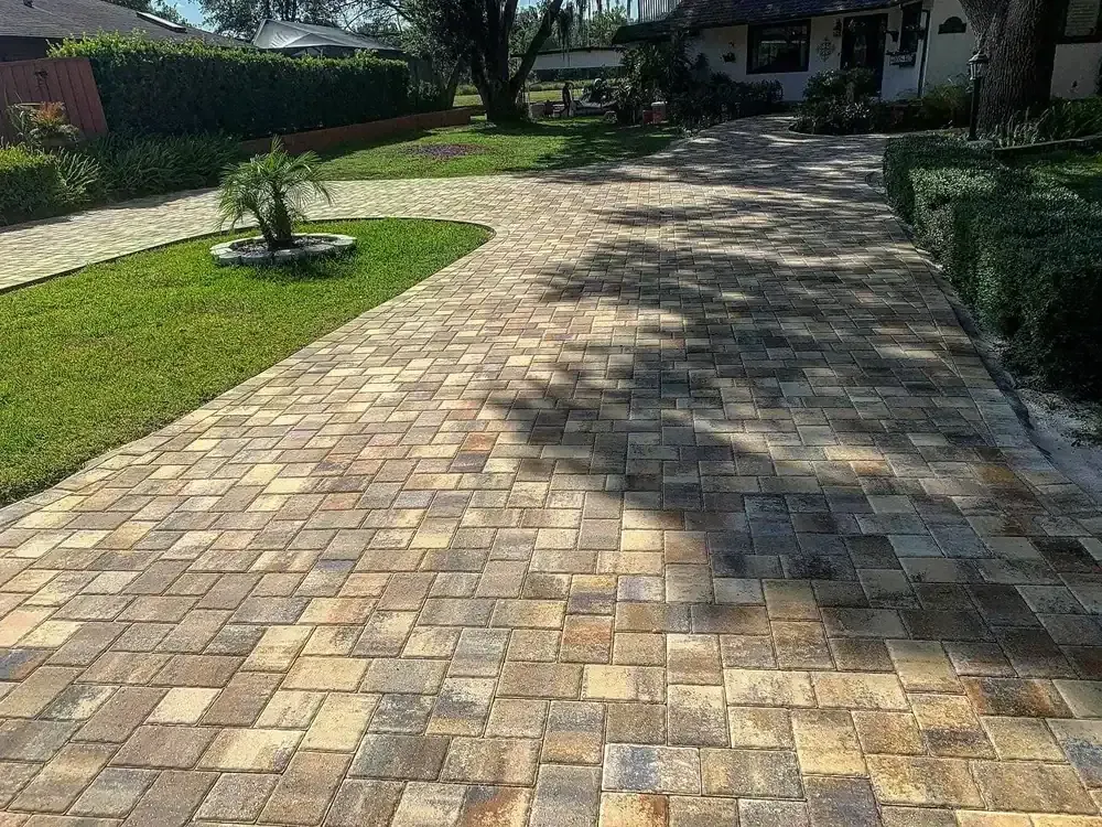 Sunlit stone-paver driveway curving past a green lawn and hedges toward a house
