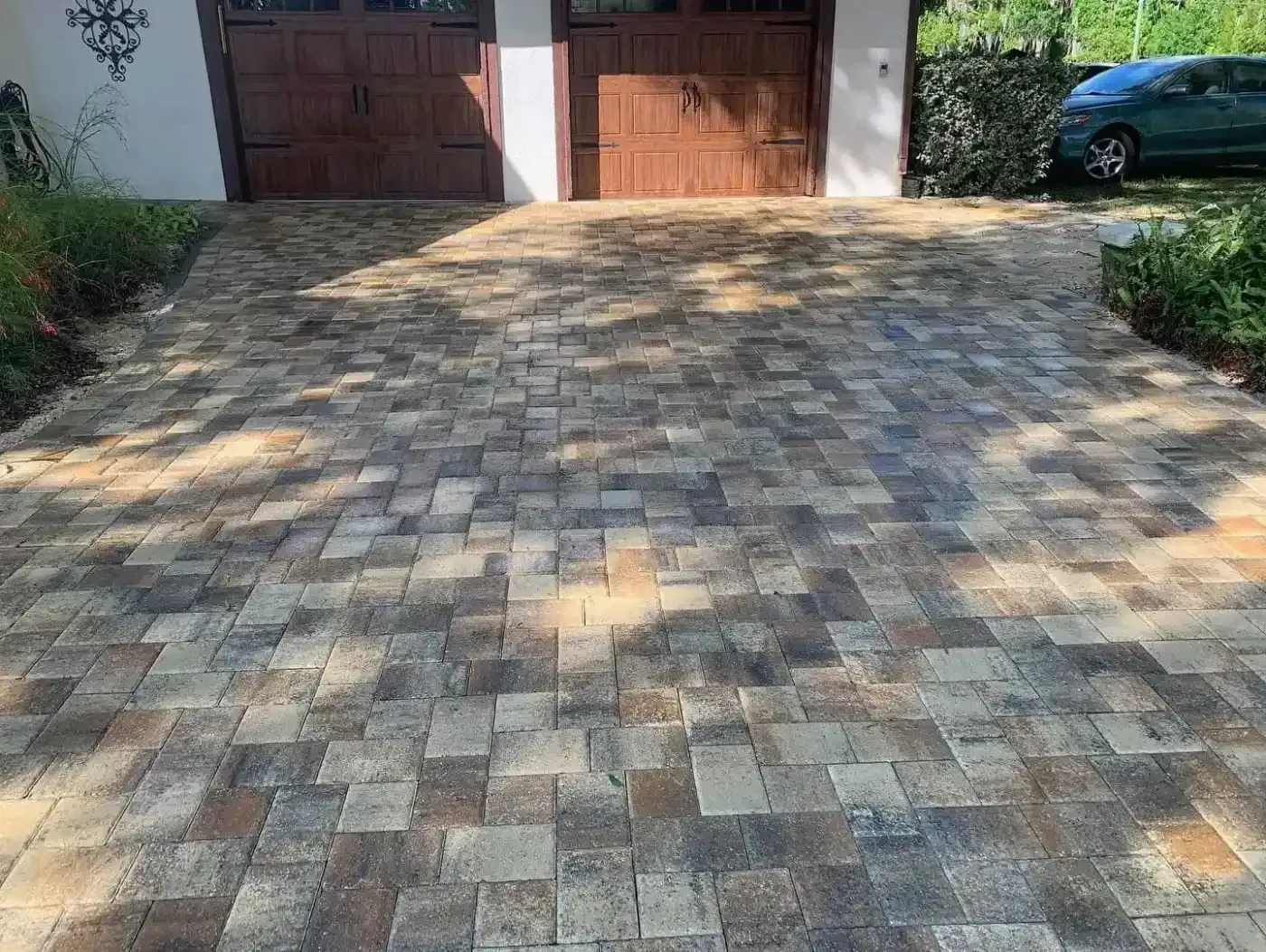 Stone driveway leading to a garage with wooden doors, bordered by greenery and a parked car on the right.