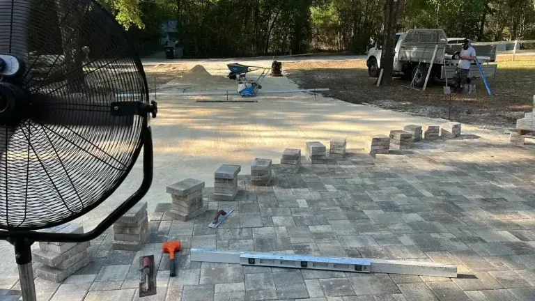 Patio paving under construction with stacked pavers, tools, and a wheelbarrow in a wooded yard
