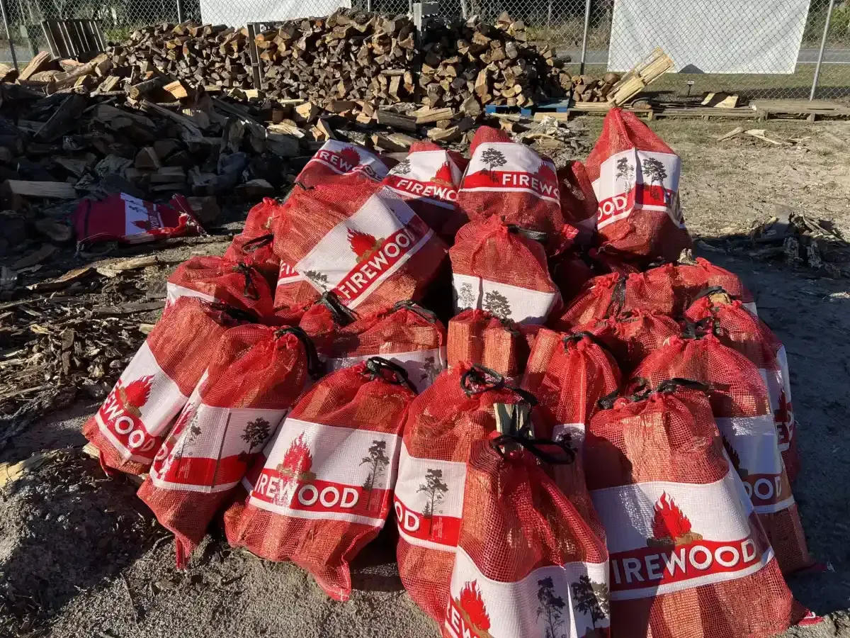 Stack of red firewood bags piled outdoors beside a woodpile and fence.