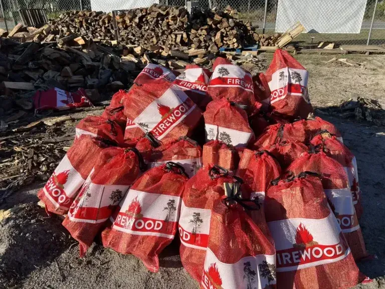 Piles of red firewood bags stacked outdoors in a yard, with loose logs in the background