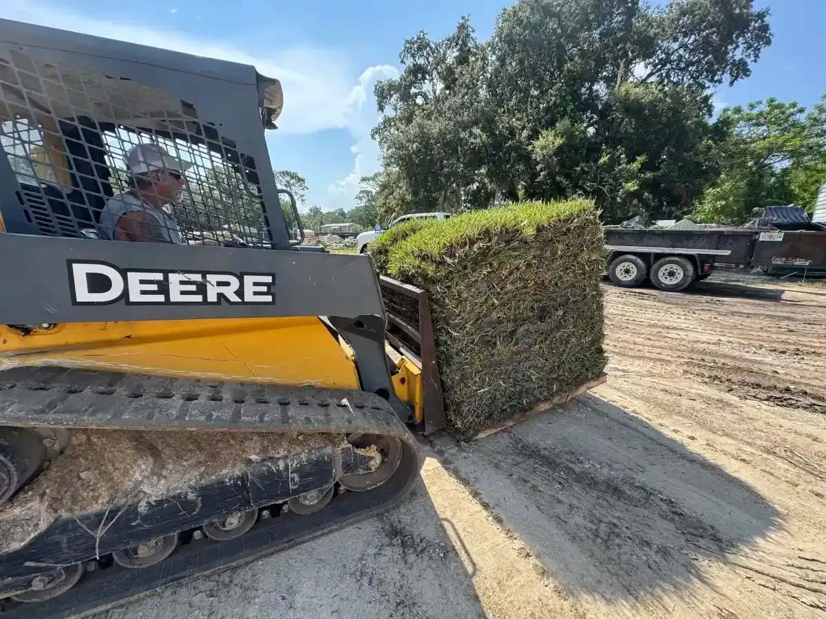 John Deere skid steer pushing a wrapped bale in a dusty field under a sunny sky