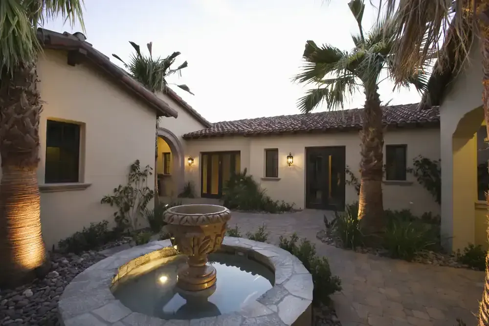 Courtyard with a central fountain, stucco buildings, palm trees, and warm exterior lighting at dusk