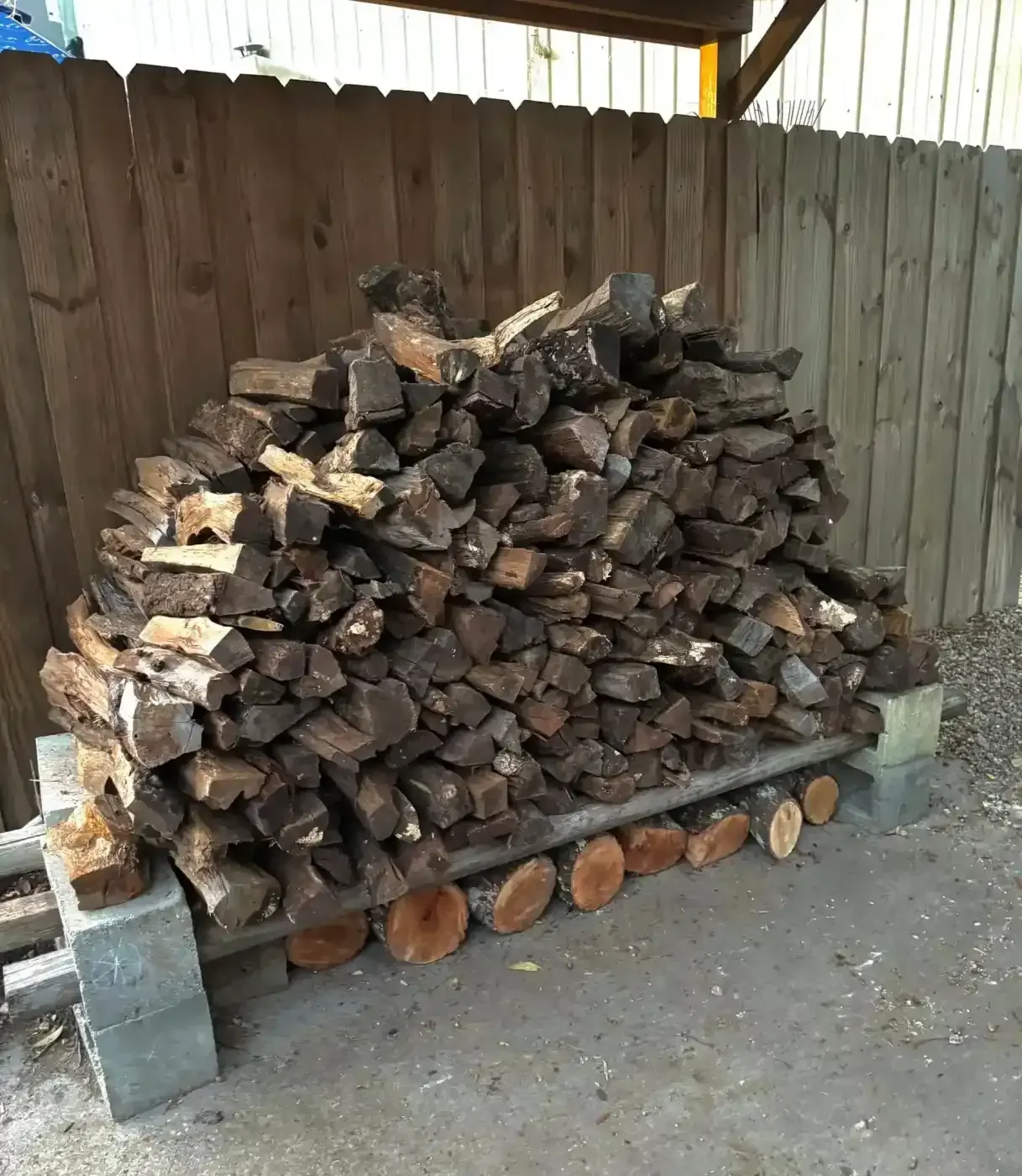 Stacked firewood under a wooden shelter beside a fence, with concrete blocks supporting the pile.