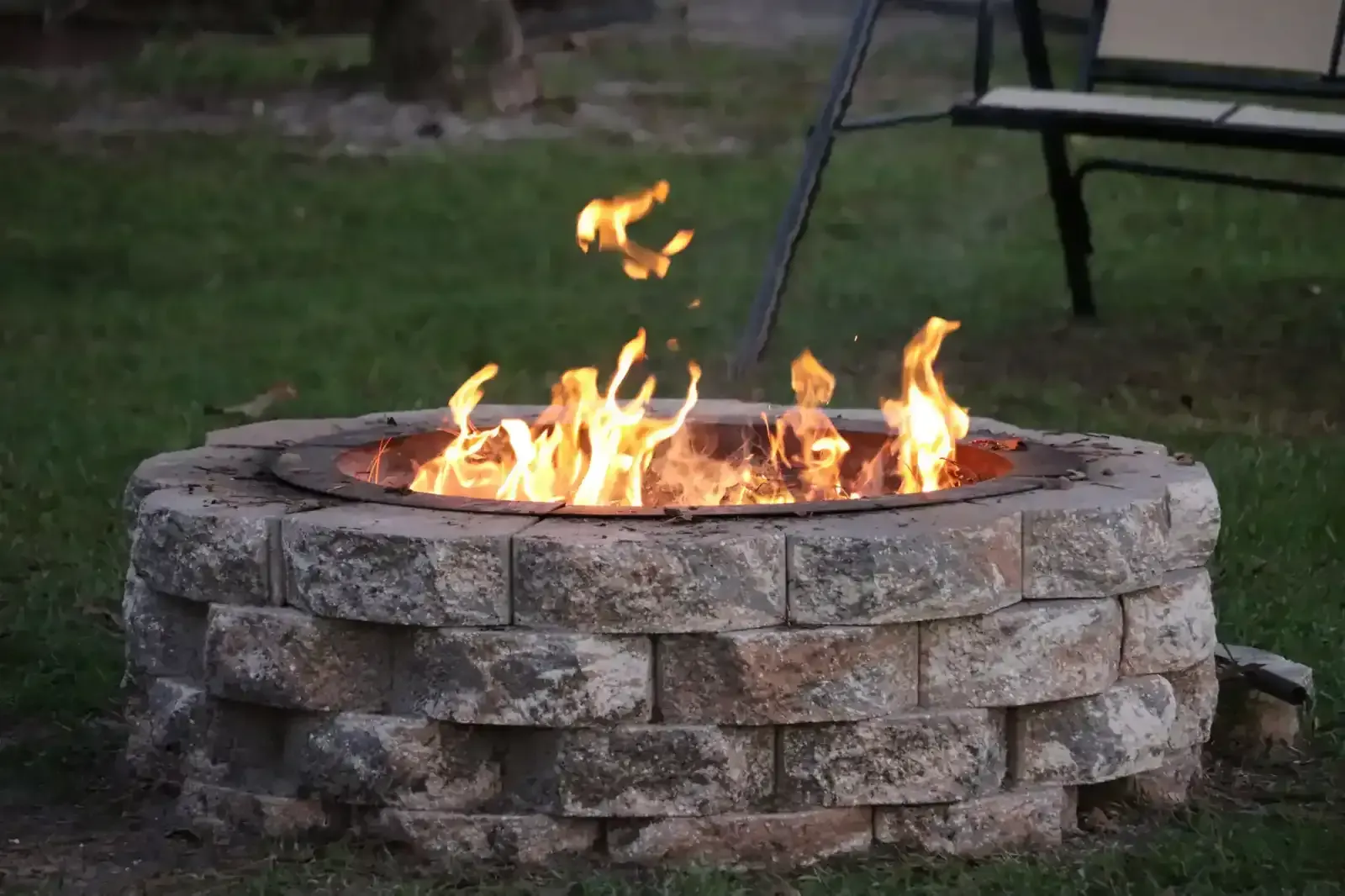 Stone fire pit with bright flames burning outdoors on a grassy lawn.