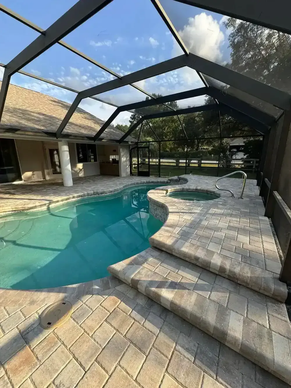 Covered backyard pool with turquoise water, stone deck, and black screened enclosure under a blue sky