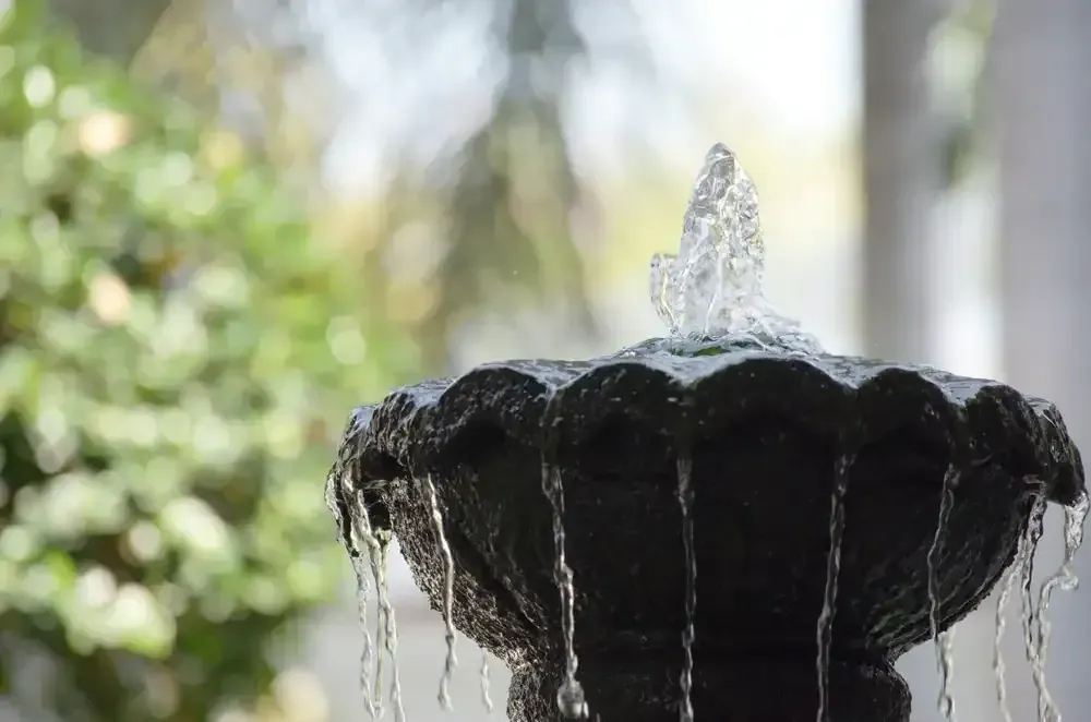 Black tiered fountain with water streaming from the top outdoors against a blurred green background