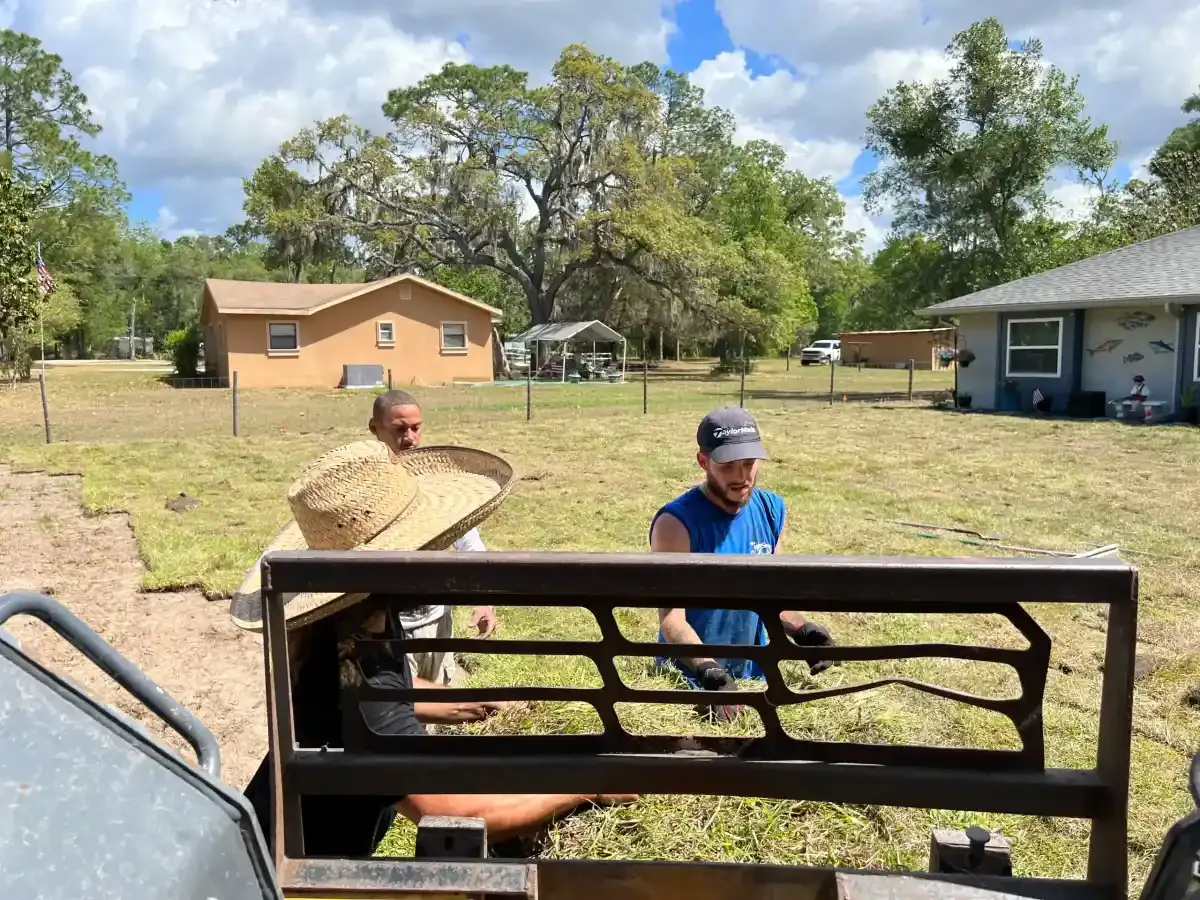 Two people working behind a metal gate in a grassy yard with houses and trees in the background