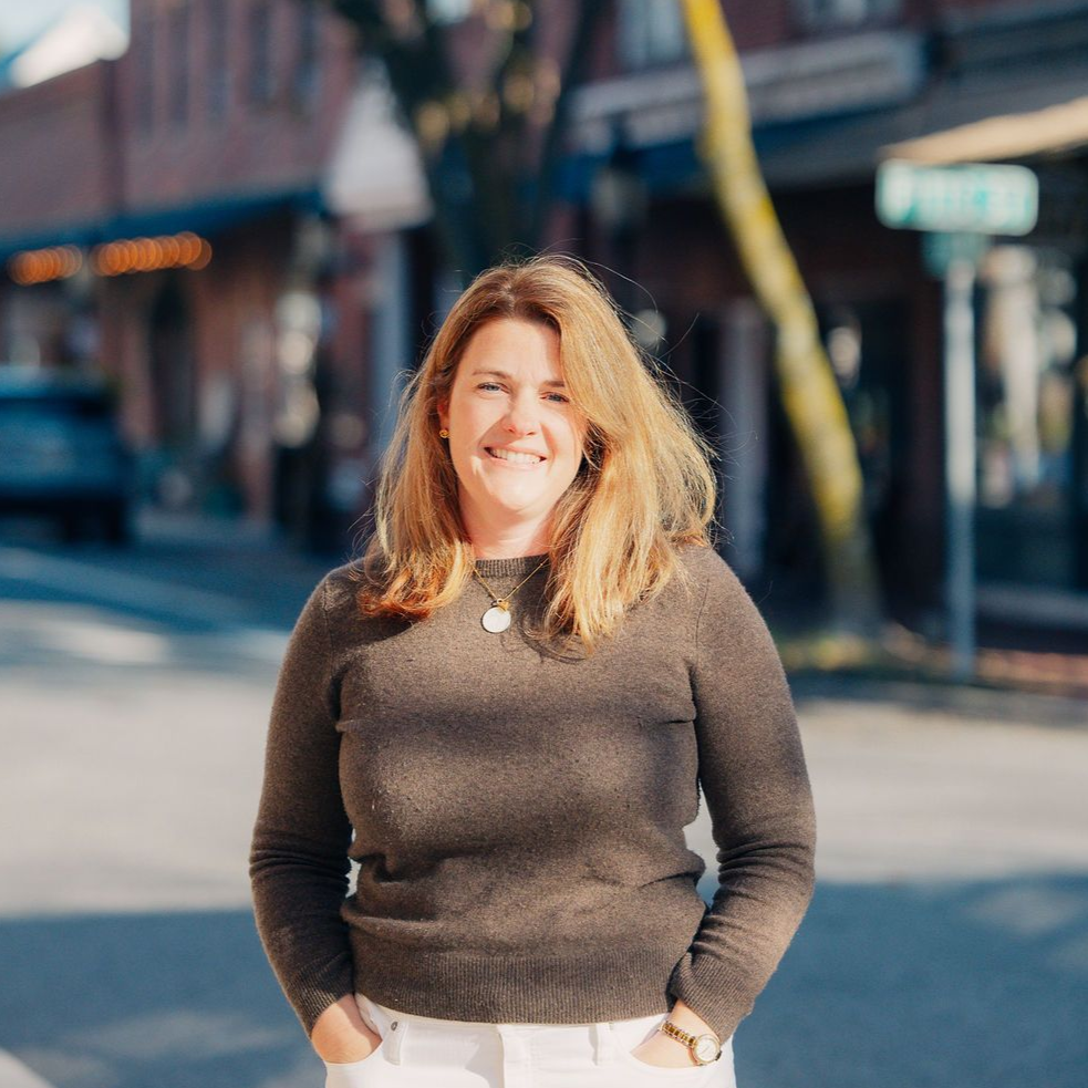 Smiling woman in a dark sweater standing outdoors on a sunny street.