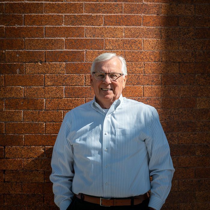 Man standing against a brick wall in a light blue shirt, smiling in sunlight