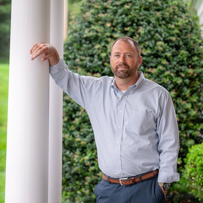 Man leaning on a white pillar outdoors, wearing a light blue shirt, with greenery in the background